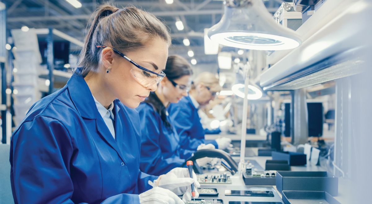 Line of women in blue workcoats working at a brightly lit production bench in an engineering factory